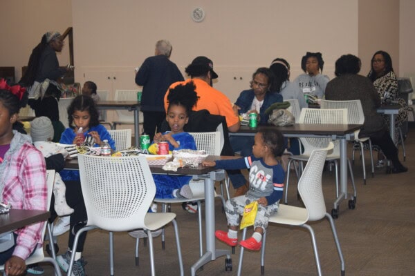 A group of people, including children and adults, are sitting at tables in a community room, eating and drinking. Some are talking, and a young child reaches for food while holding a snack bag.