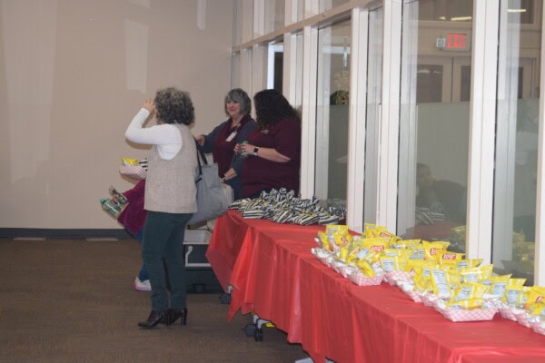 Three women stand near a wall with windows, next to tables covered in red tablecloths and filled with bags of chips and snacks. One woman faces away, while the others are engaged in conversation.
