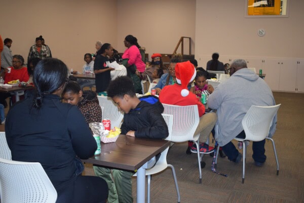 A group of people, including children and adults, sit at tables in a community room eating food. One person is wearing a red Santa hat. The atmosphere appears friendly and social.