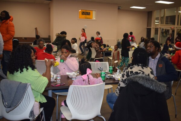 A group of people, including adults and children, sit at tables eating and drinking in a large, brightly lit room. More people are seen mingling and sitting at other tables in the background.