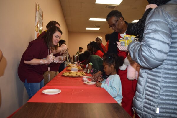 A woman wearing gloves serves cookies to children and adults lined up at a long table with a red tablecloth in a community setting. The room is busy, and people are engaged in the activity.
