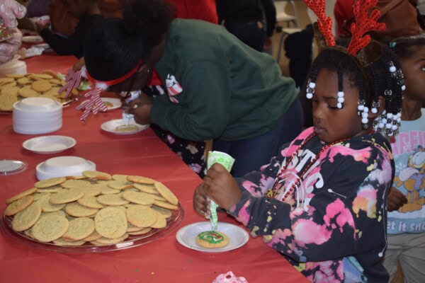 A young girl decorates a cookie with icing at a table covered with sugar cookies. She wears beaded hair and reindeer antlers. Other children are also decorating cookies in the festive setting.