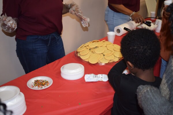 A child decorates cookies at a table covered with a red cloth. Plates of plain cookies, sprinkles, and paper plates are on the table. Two adults wearing gloves are helping with the activity.