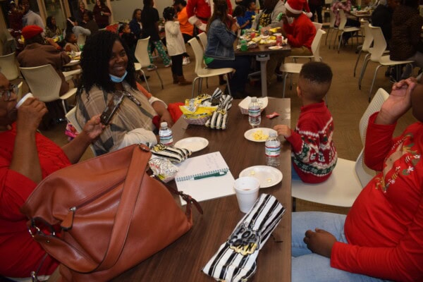 People sit around a table at a holiday gathering, eating snacks. A woman smiles, a child in a festive sweater sits beside an adult, and a large brown purse is on the table. Other groups are visible in the background.