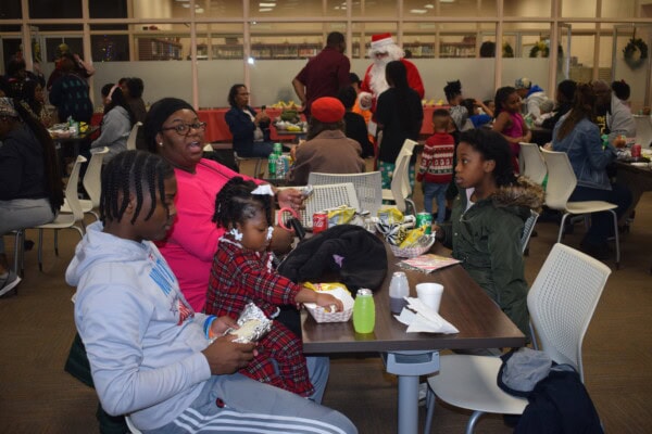 Families and children sit at tables eating snacks in a crowded room. A woman and children are in the foreground, while a person dressed as Santa Claus stands in the background. People are talking and enjoying the event.