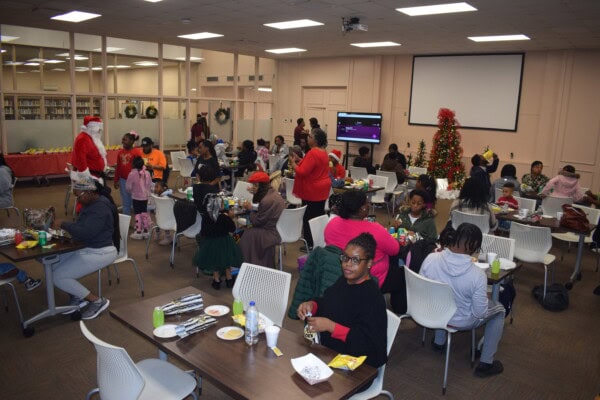 A large group of people, including children and adults, sit at tables in a decorated room with a Christmas tree and Santa Claus present. The scene is festive, with food and drinks on the tables and holiday decorations visible.