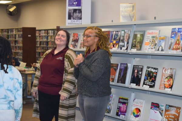 Two women stand and smile in a library next to a magazine display. Bookshelves and more magazines are visible in the background. One woman is clapping, and the other has her hands by her sides.