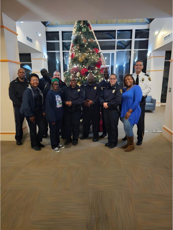A group of people, some in police uniforms and others in casual or business attire, stand smiling in front of a large decorated Christmas tree inside a building with tall windows.