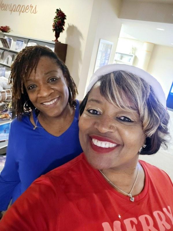 Two women smiling for a selfie indoors. The woman in front wears a Santa hat and a red shirt with MERRY on it; the woman behind has shoulder-length hair and a blue top. They appear happy and festive.