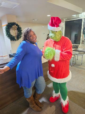 A woman in a blue dress and brown boots poses and smiles next to a person dressed as the Grinch in a Santa costume, standing indoors by a reception area with a holiday wreath on the wall.