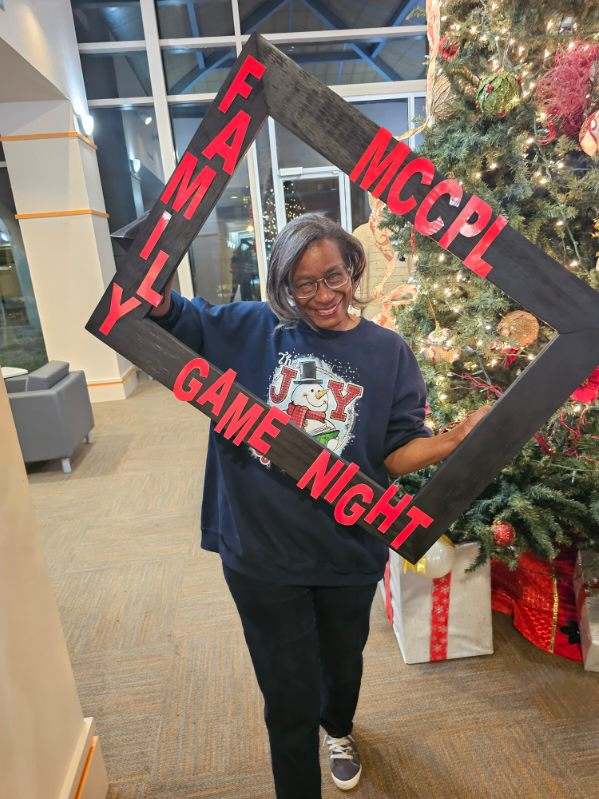 A woman smiles while holding a large frame with red letters reading MCCPL Family Game Night in front of a decorated Christmas tree indoors. She wears glasses and a dark Joy holiday sweater.