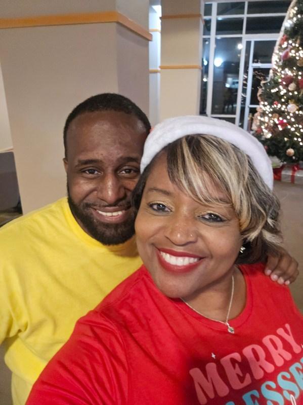 A smiling woman in a Santa hat and Merry Blessed shirt takes a selfie with a smiling man in a yellow shirt. A decorated Christmas tree is visible in the background.