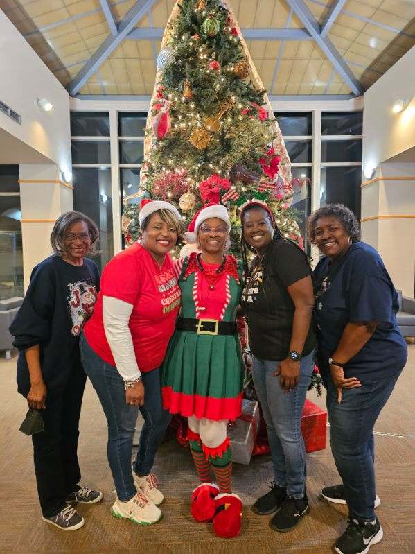 Five smiling women pose in front of a decorated Christmas tree. The center woman is dressed as an elf, and the others wear festive shirts. Wrapped presents are visible under the tree in a warmly lit room.