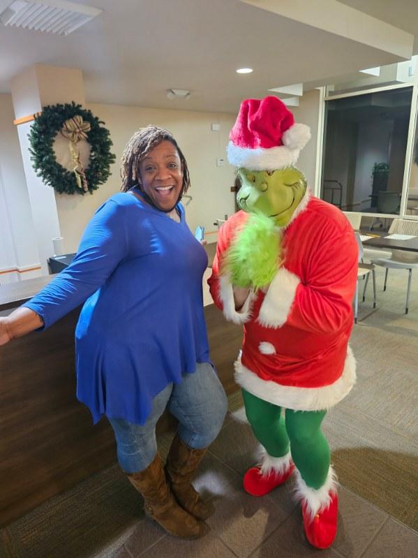 A smiling woman in a blue tunic and brown boots poses next to someone dressed as the Grinch in a Santa outfit inside a decorated, indoor setting.
