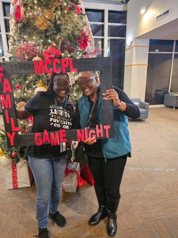 Two women smile and pose together inside a large photo frame labeled “MCCPL Family Game Night” in front of a decorated Christmas tree with wrapped gifts underneath in a festive indoor setting.