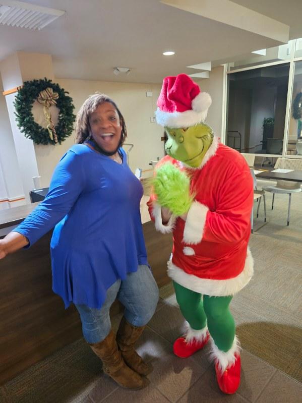 A woman in a blue top and brown boots poses happily next to a person dressed as the Grinch in a Santa outfit, inside a room decorated with holiday wreaths.