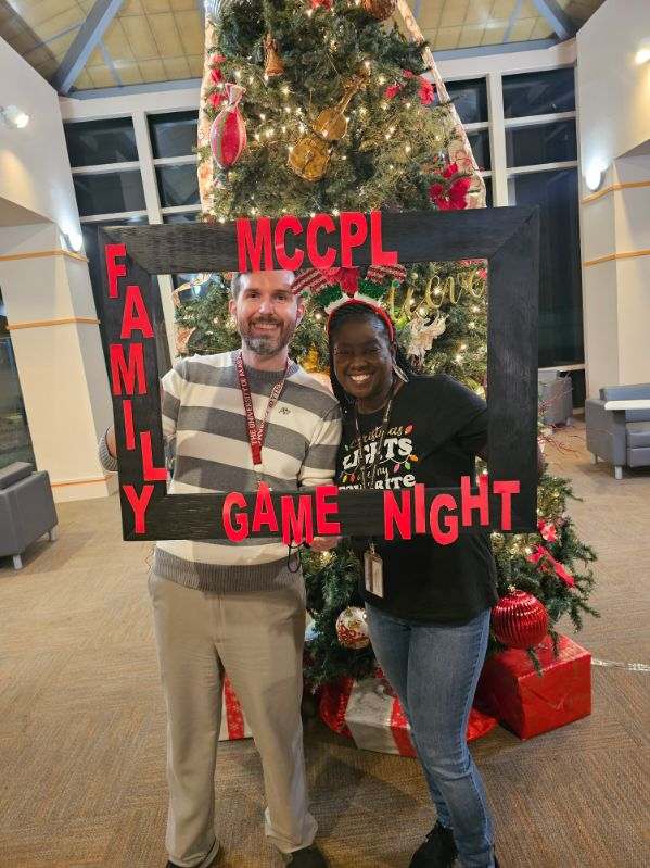 Two people smile while holding a large black frame that says MCCPL Family Game Night in front of a decorated Christmas tree inside a festive, well-lit room.