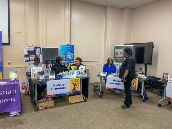 A group of people at a book fair with tables displaying books, signs for Katrice Crawford and Willie Freeman Jr., and promotional materials in a brightly lit room. One person stands, while others sit behind the tables.