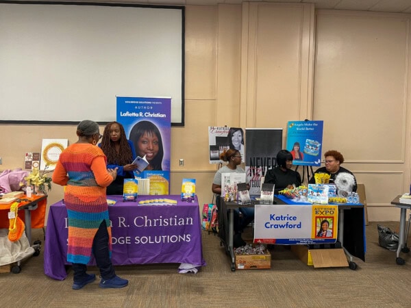 Three women sit behind tables displaying books, signs, and promotional materials at an indoor event. Another woman stands, interacting with them. Colorful banners and book covers are prominently featured on the tables.