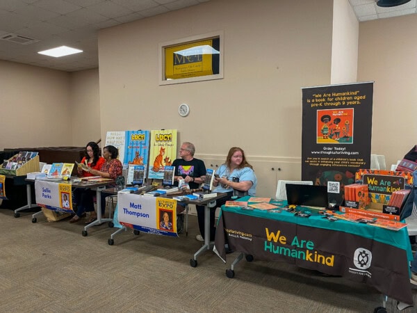 Four authors sit behind tables displaying their books at a book fair. Each table features signs with the authors’ names and books. A large banner on the right promotes We Are Humankind. Books and promotional materials are visible.