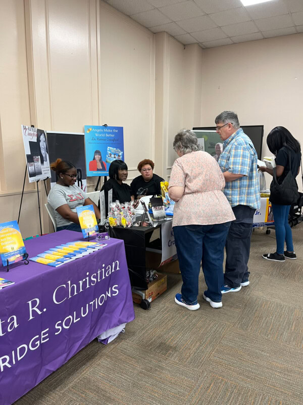 People gather around a table with books, pamphlets, and gift items at an indoor event. A purple tablecloth reads Rita R. Christian BRIDGE SOLUTIONS. Two women sit behind the table, talking with visitors.