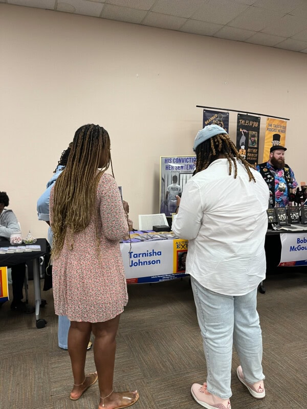 Two people stand facing a table at a book fair, looking at displays and books. The table has a sign with the name Tarnisha Johnson. Other tables with banners and books are visible in the background.