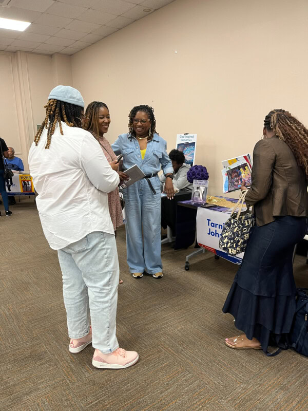 Four women are standing and talking at an indoor event with tables and displays in the background. Two women are holding notebooks, and one is wearing a blue jumpsuit. A woman sits behind a table with promotional materials.