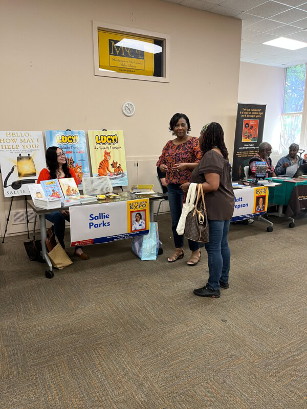 Two women stand and talk in front of a table displaying childrens books and a Sallie Parks sign at an indoor event. Other authors and tables with books are visible in the background.