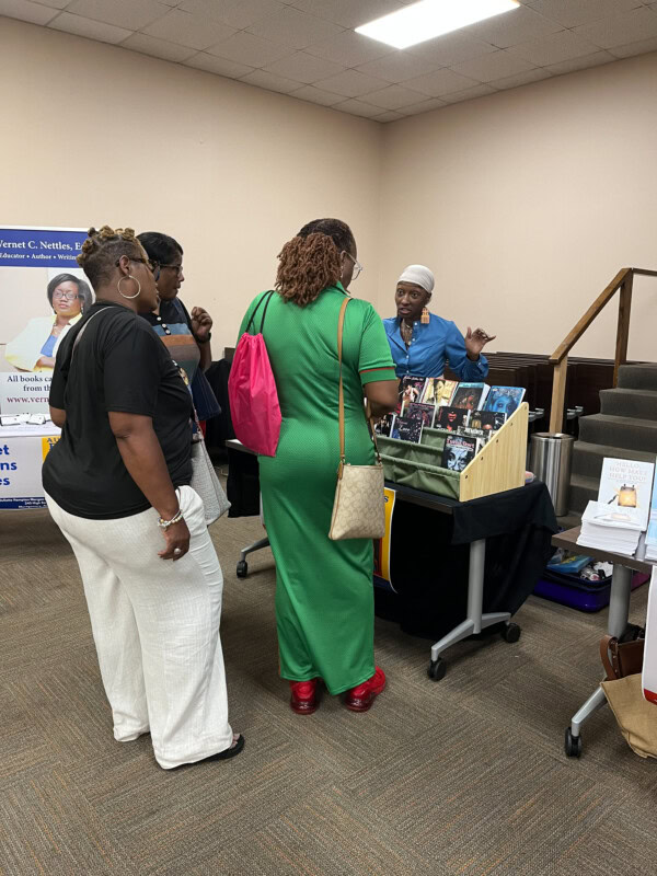 Three women stand at a vendor table, engaging with a person behind the table who is displaying books and photos. The setting appears to be an indoor event or fair with informational displays.