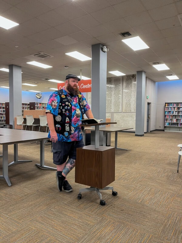 A person with tattoos, a beard, and colorful clothing stands at a podium in a library, speaking to an unseen audience. Bookshelves and empty chairs are visible in the background.