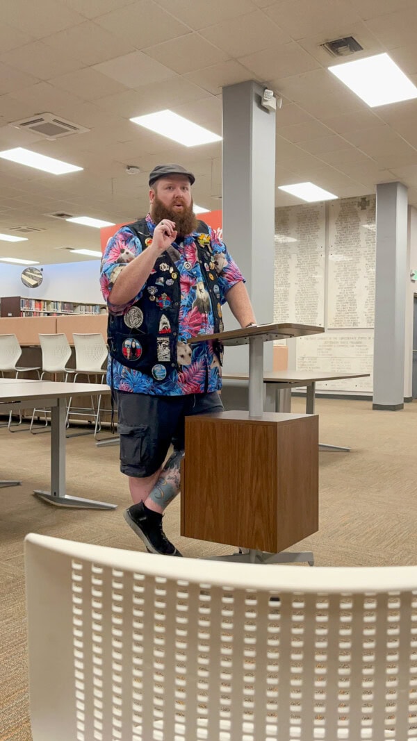 A bearded person in a colorful shirt, decorated vest, shorts, and cap speaks at a podium in a library or classroom, with white chairs and tables arranged in the background.