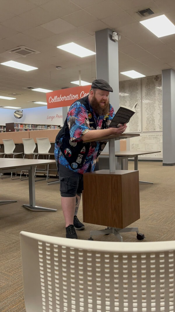 A bearded person wearing a colorful shirt, black shorts, and a cap leans on a podium while reading a book aloud in a library’s “Collaboration Center.” Empty chairs and tables are visible in the background.