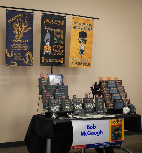 A display table at a book expo features books, promotional materials, and banners for “Tales by Bob.” The table has stacks of books, decorations, and a sign reading “Bob McGough.” Banners hang behind, promoting the author and podcast.