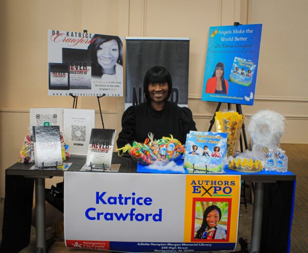 A woman sits at a table displaying books, flyers, and promotional materials at an expo. A large sign reads Katrice Crawford, with photos of her books and a poster about her author event.
