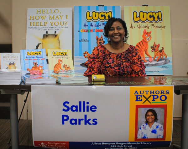A smiling woman sits at a table displaying her childrens books about a cat named Lucy at an Authors Expo. A sign reads Sallie Parks and colorful book covers are shown behind her.