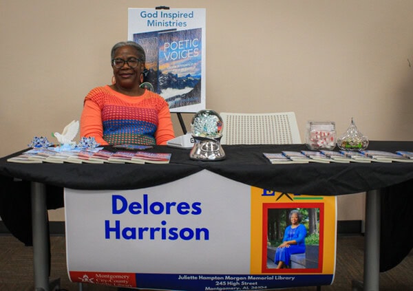 A woman with short gray hair and glasses sits at a table with books, brochures, and a glass ornament. A banner with Delores Harrison and her photo hangs in front. A Poetic Voices poster is displayed behind her.