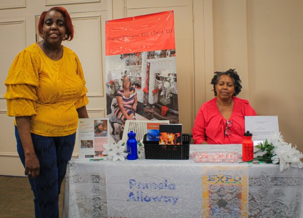 Two women are at a table with promotional materials and a banner behind them. One woman stands smiling in a yellow blouse; the other sits in a red top. The table displays flyers, water bottles, and decorative items.
