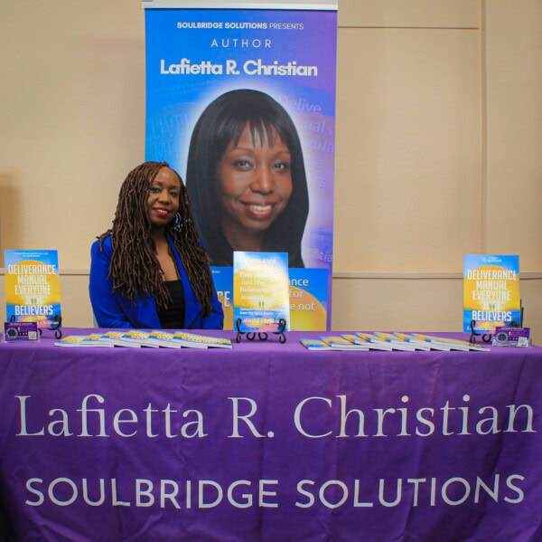 A woman with long braids sits at a table covered with a purple cloth that reads “Lafietta R. Christian, Soulbridge Solutions.” Behind her is a banner with her photo and books arranged on the table.