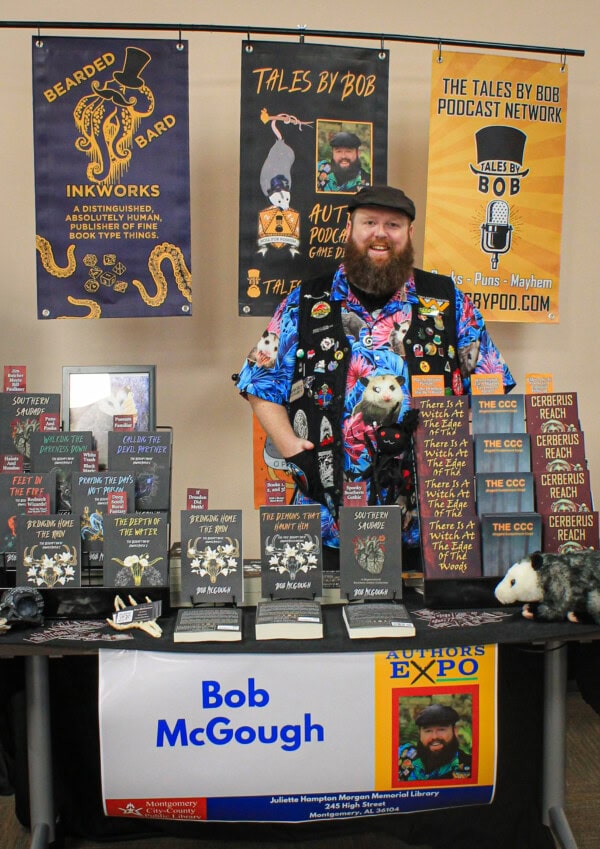 Bob McGough, a bearded man in a colorful shirt, stands behind a table displaying his books and podcast signs at an expo. The table bears his name and is decorated with banners, stuffed animals, and book displays.