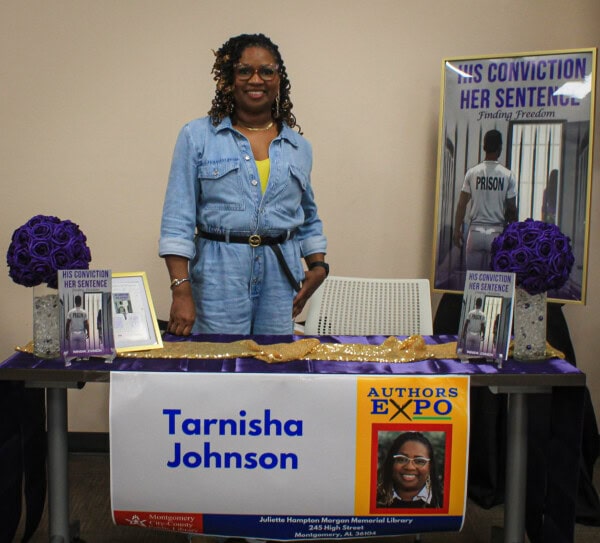 A woman in a blue denim dress stands behind a table displaying books, flowers, and a sign that reads Tarnisha Johnson, Authors Expo. A large poster of her book, “His Conviction, Her Sentence,” is in the background.