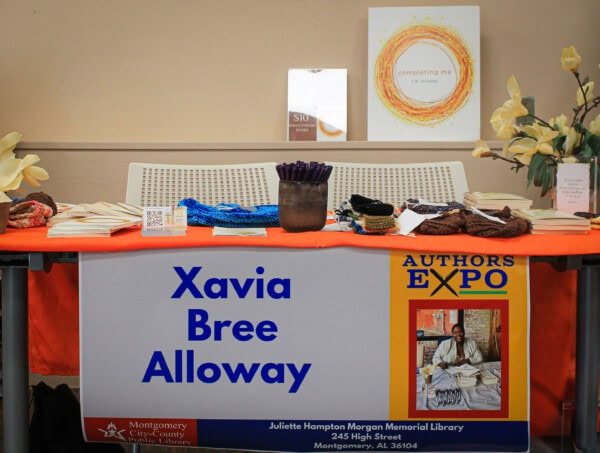A table with an orange cloth displays books, pens, and accessories. A large sign reads Xavia Bree Alloway and Authors Expo at Juliette Hampton Morgan Memorial Library. A framed photo and posters are in the background.