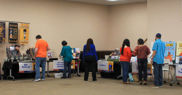 People browsing and talking to authors at tables in a book fair; each table displays books and signs with author names, set up in a large indoor room with posters on the walls.