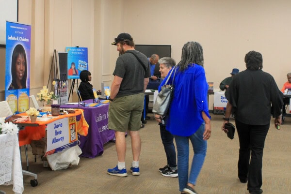 People browse informational tables at an indoor community event. Various displays with banners, brochures, and items are set up, and attendees are walking and talking with exhibitors.