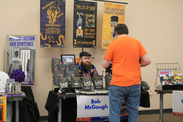 A man with a beard and a hat sits at a table covered with books and promotional materials, labeled “Bob McGough.” Another man in an orange shirt stands in front of the table. Colorful posters hang behind the author.