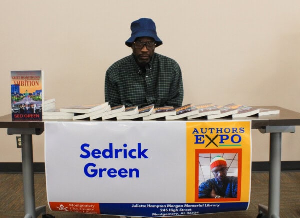 A man in a blue hat and glasses sits behind a table with books and a sign that reads Sedrick Green and Authors Expo at a library event. One of his books, titled Ambition, is displayed upright on the table.