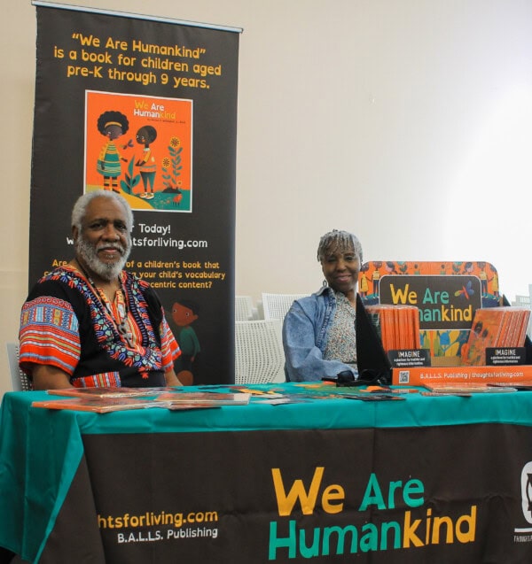 Two authors sit at a table covered with We Are Humankind promotional materials and books. Behind them is a banner showing the book cover and information. Both are smiling and facing the camera.