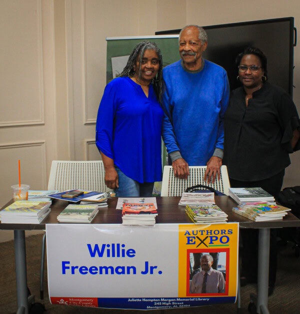 Three people stand behind a table with books and a sign reading Willie Freeman Jr. Authors Expo. The table is set up in a library or event space, with pamphlets, books, and a drink on it.