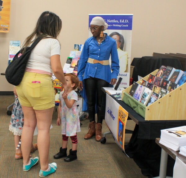 A woman and two young girls talk to a vendor at a book fair booth, where books and brochures are displayed. The vendor stands behind the table, wearing a blue shirt and tan headwrap.