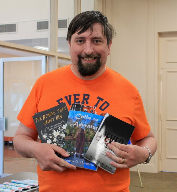 A man in an orange shirt stands indoors, smiling and holding three books titled “The Demons That Haunt Him,” “Calls to Adventure,” and “Severed.”.