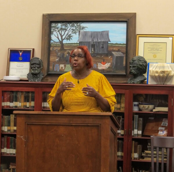 A woman with short red hair, wearing a yellow blouse, speaks at a podium in a library. Behind her are books, busts, and framed awards, with a painting of a farmhouse hanging on the wall.